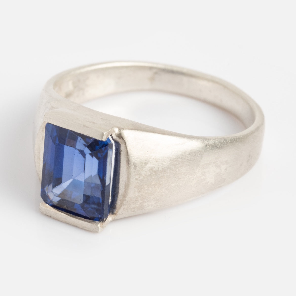 The Legacy Ring is a silver band with a rectangular, faceted blue gemstone, showcased against a plain white background.