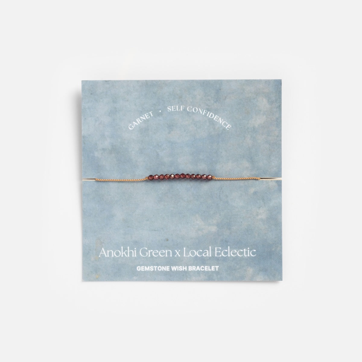 The Anokhi Green x Local Eclectic Gemstone Wish Bracelet, featuring delicate natural garnet beads for self-confidence, is shown on a blue card labeled with the bracelet name and "Garnet • Self Confidence.