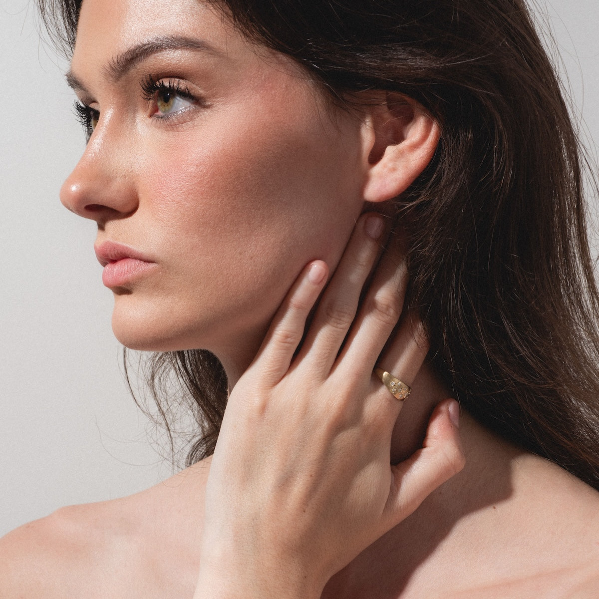 A woman with long brown hair poses in profile against a light background, gently touching her neck and showcasing the Half Moon Bombe Ring on her finger, complemented by a natural, minimal makeup look.