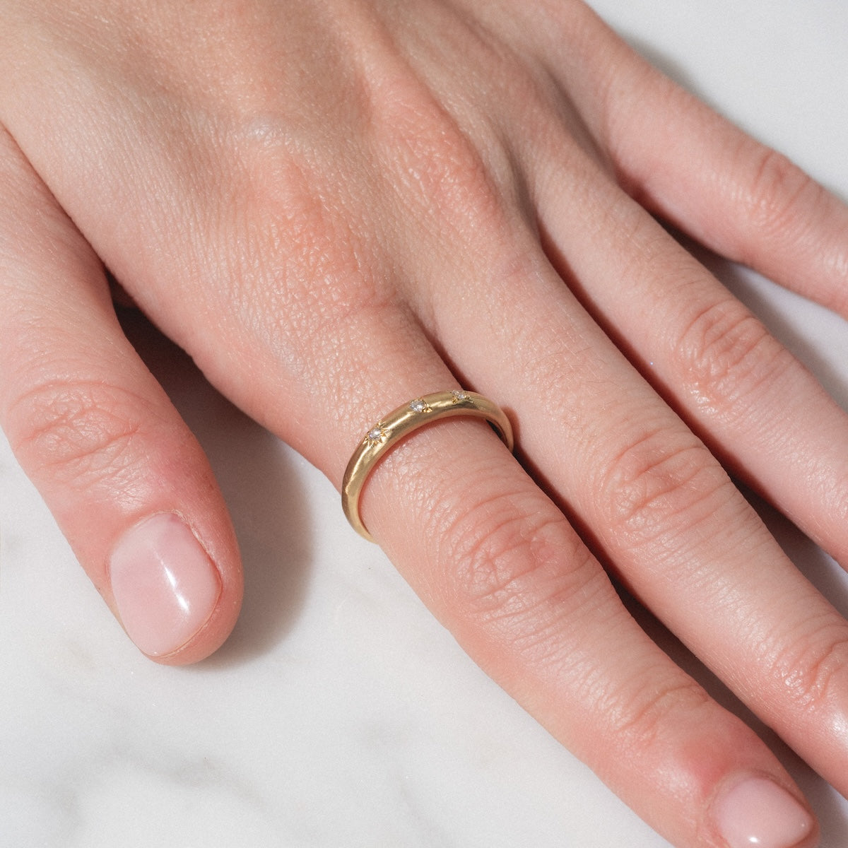 A close-up of a hand with neatly trimmed nails wearing the "You are my Sun, Moon and Star Band" gold ring on the ring finger, resting on a white marble surface.