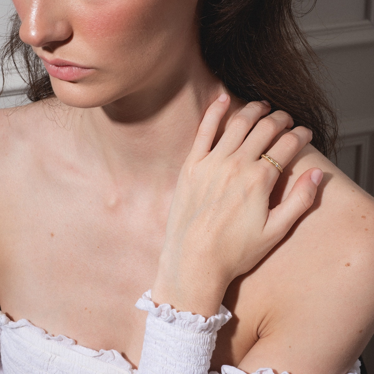 A woman with wavy brown hair and natural makeup rests her hand on her collarbone, showcasing the "You are my Sun, Moon and Star Band" gold ring. She wears a white off-shoulder top, and her face is partially visible.