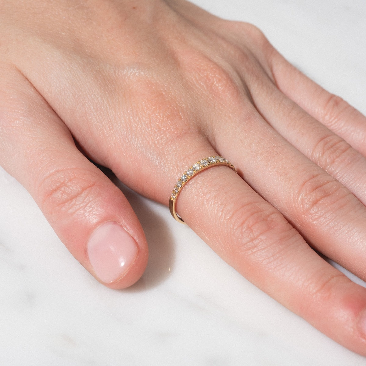 A close-up of a hand with natural, neatly trimmed nails rests on a white surface, showcasing the Echo Diamond Band—a thin gold ring with small diamonds on the ring finger.