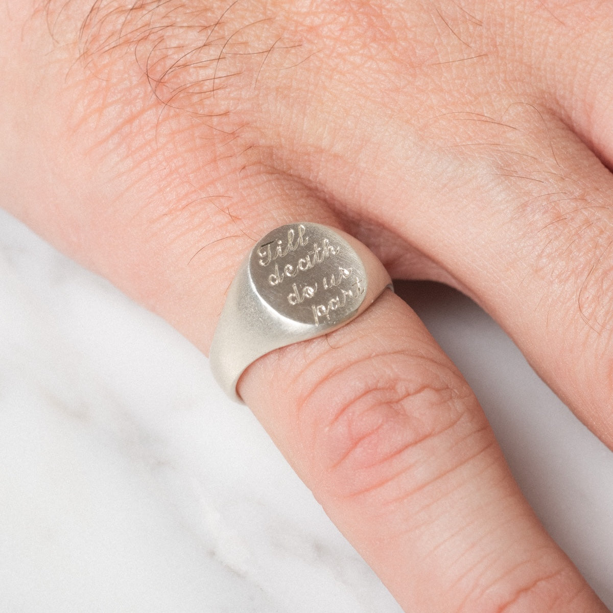 A close-up of a hand wearing the Till Death Do Us Part Signet Ring, engraved with the iconic phrase, resting on a white marble surface.
