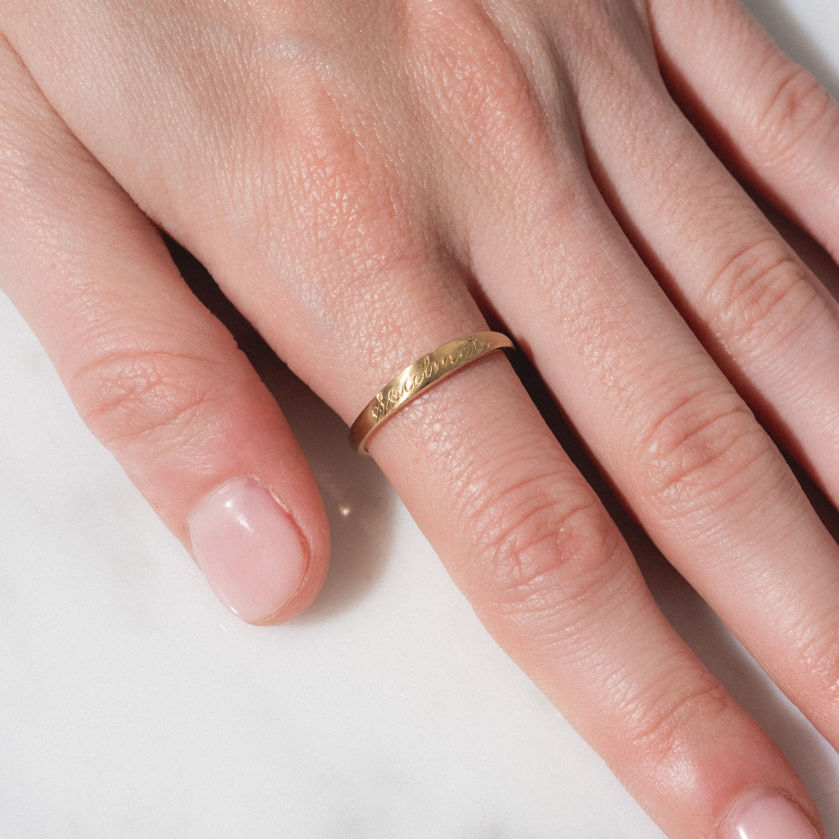 A close-up of a hand with neatly manicured nails wearing the Soulmate Band, a simple gold ring, on the ring finger, resting on a white marble surface.