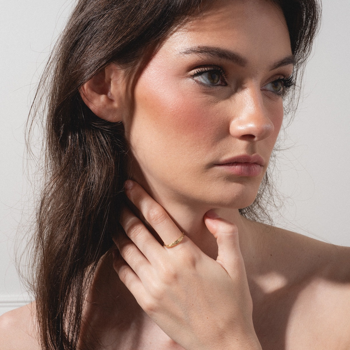 A woman with long brown hair and natural makeup looks to the side, softly touching her neck to display the Soulmate Band—a simple gold ring—on her finger. Gentle light highlights her face and bare shoulders.