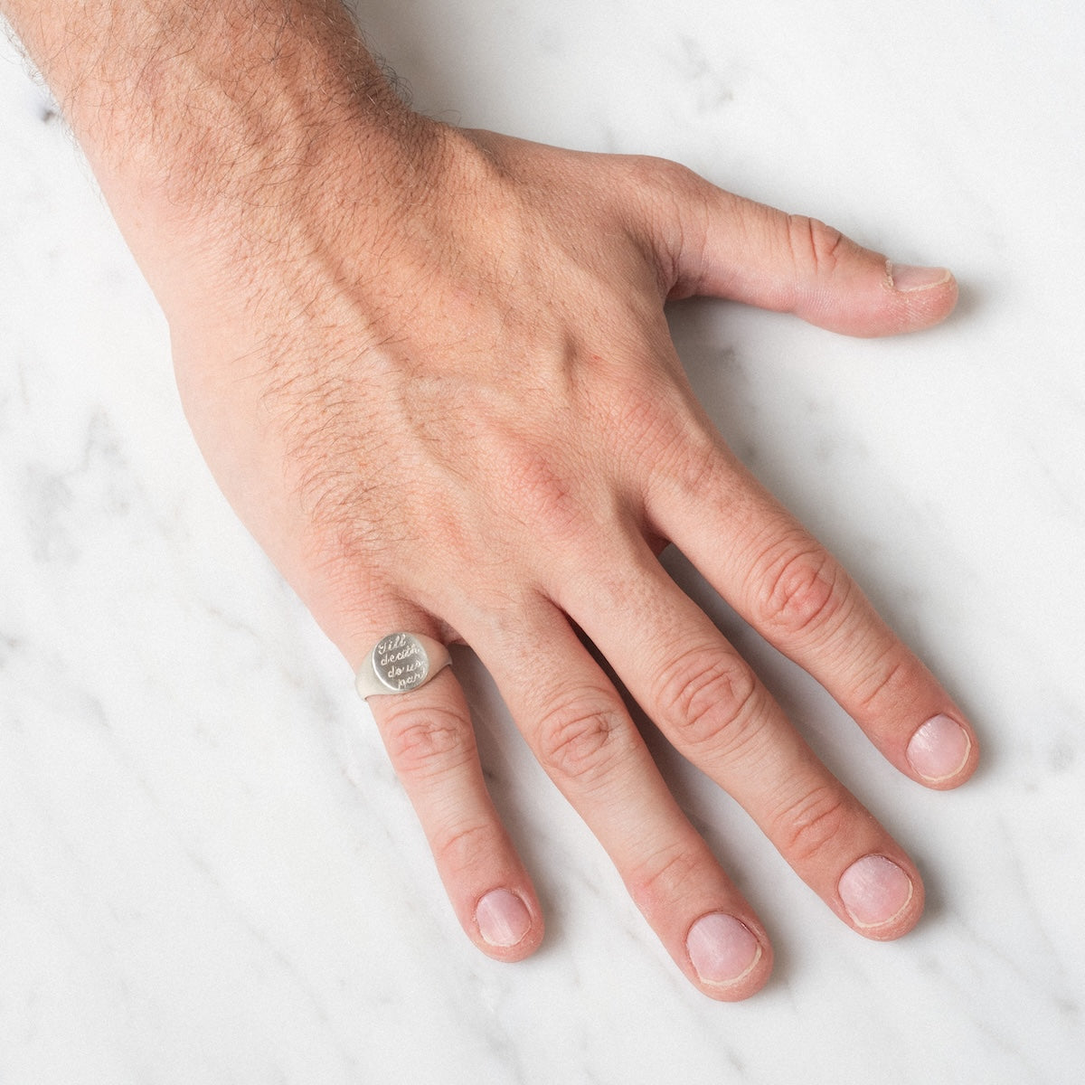 A left hand rests on white marble, wearing the Till Death Do Us Part Signet Ring with engraved text on the pinky finger.