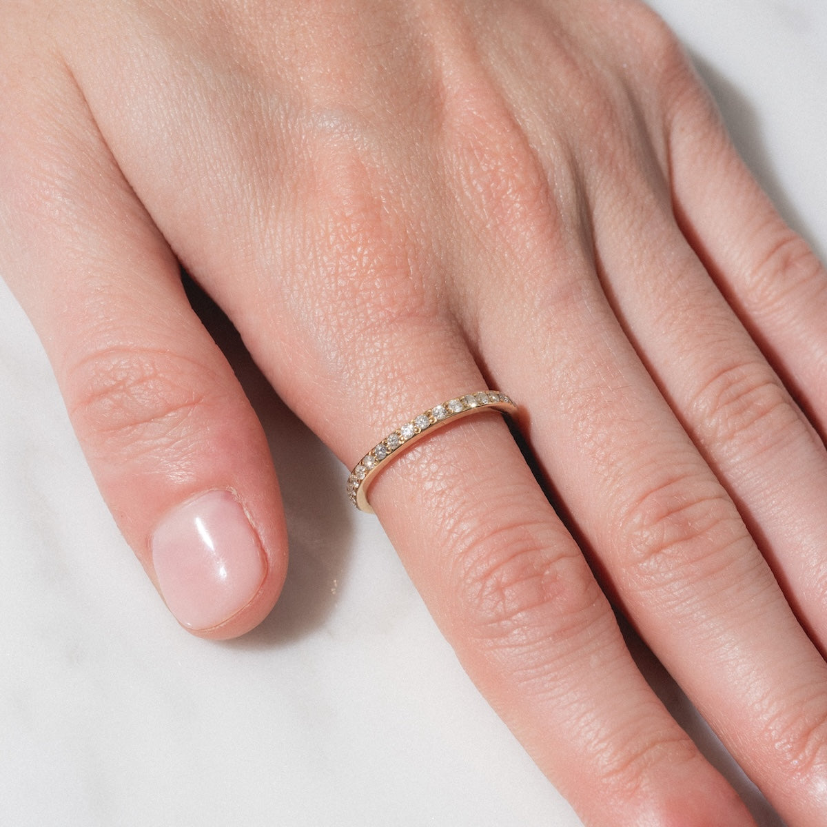 A close-up of a hand on a white surface showcases the Infinity Diamond Band—a thin gold ring with small diamonds on the ring finger. The nails are neatly trimmed and natural.
