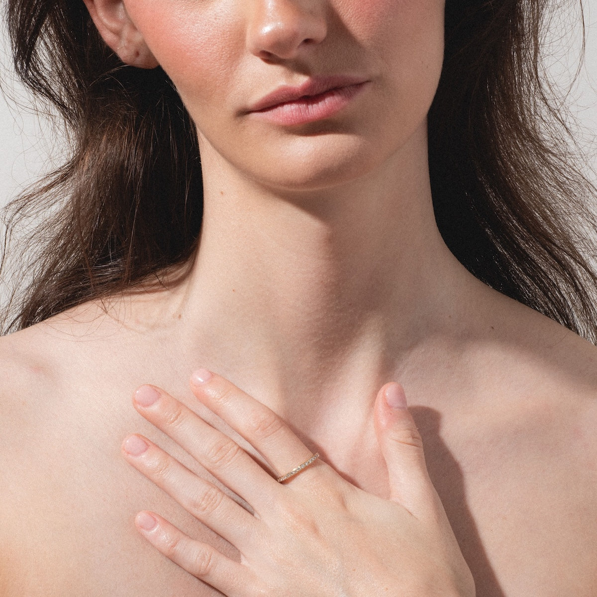 A woman with long brown hair touches her chest with her left hand, displaying the Infinity Diamond Band—a delicate gold ring accented with small diamonds on her ring finger. She stands against a simple, light background.
