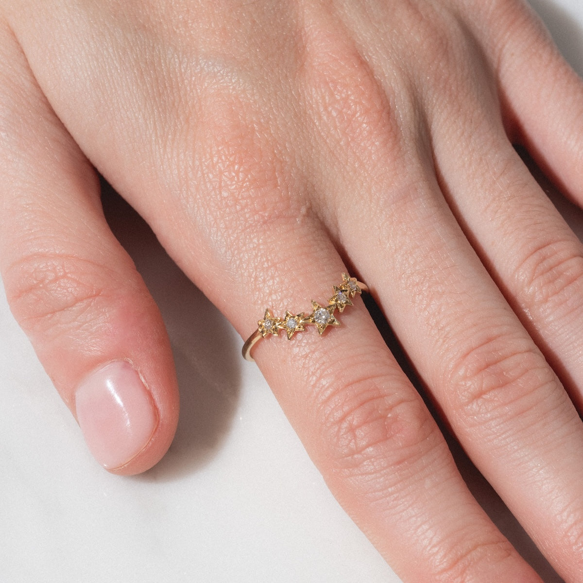 A hand wearing the Stars Aligned Band—a gold ring adorned with sparkling, star-shaped designs—rests on a white surface, the ring gracing the ring finger.