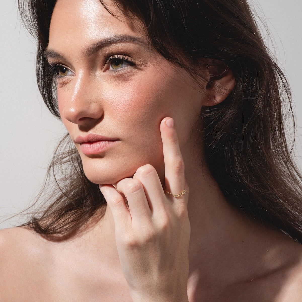 A woman with long brown hair gazes thoughtfully to the side, her hand gently touching her cheek. She wears the Stars Aligned Band, a delicate gold ring, and her natural makeup glows softly under gentle lighting.