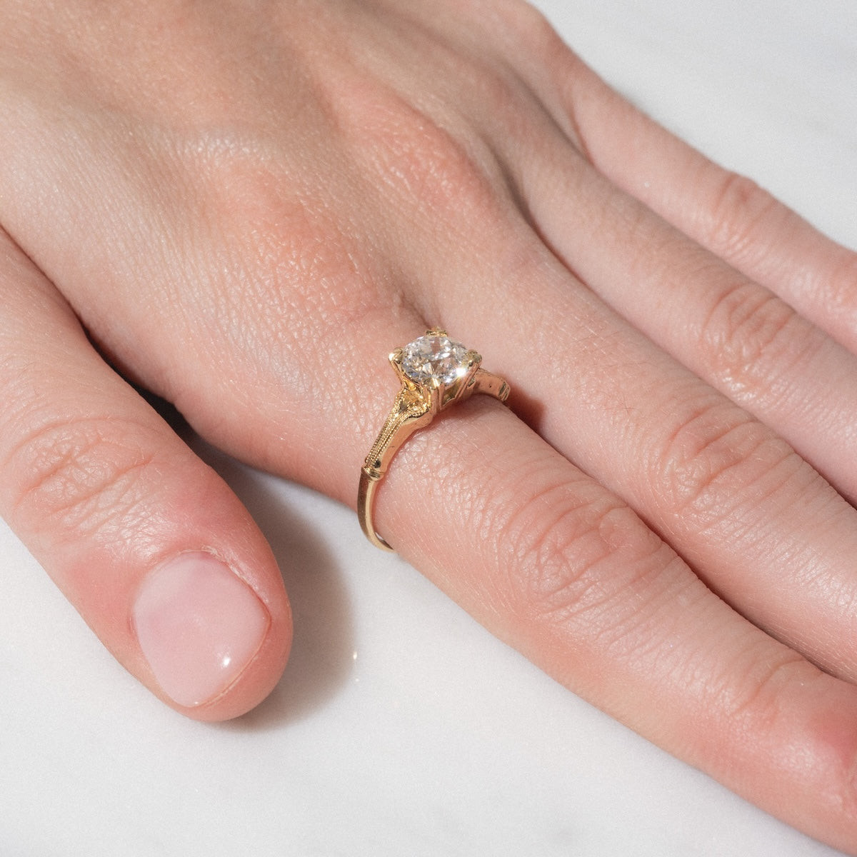 A close-up of a hand with natural, unpolished nails resting on a white surface, wearing the Elara Ring—a gold band featuring a large, round diamond on the ring finger.