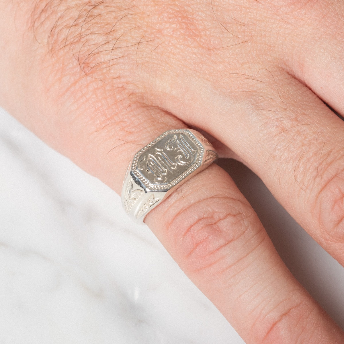 A close-up of a hand wearing the Sovereign Signet Ring, a silver piece with intricate engravings and Arabic script on its rectangular top, resting on a white marble surface.