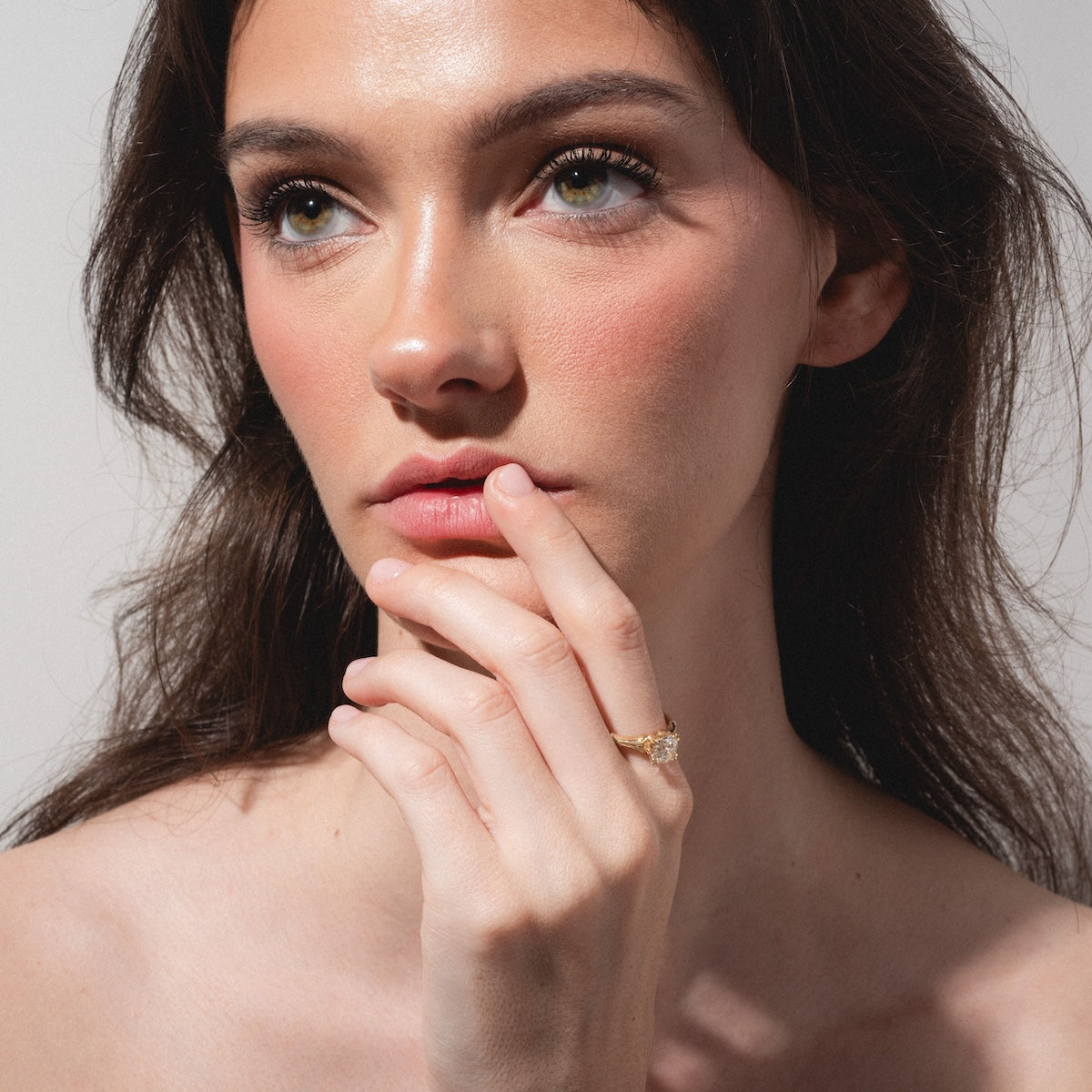 A woman with long brown hair and natural makeup gazes thoughtfully to the side, her hand with the Elara Ring near her lips. She wears the elegant gold band on her finger against a softly lit, plain background.