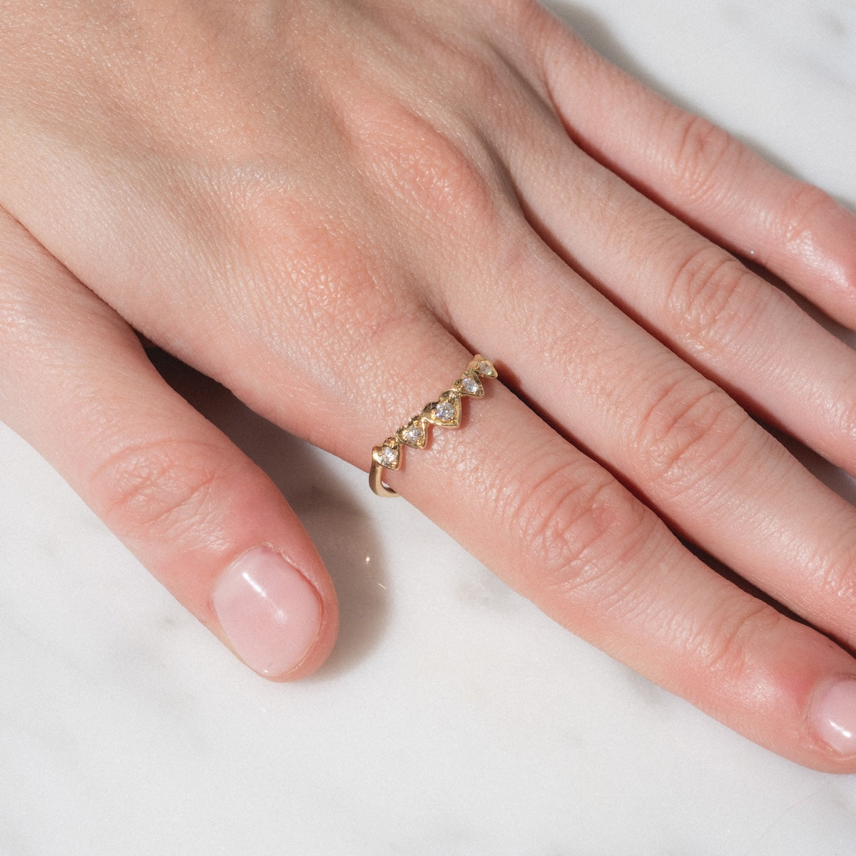 A close-up of a hand with clean, natural nails rests on a white surface, wearing the Radiant Hearts Band (outward band), a gold ring adorned with five heart-shaped diamonds on the ring finger.