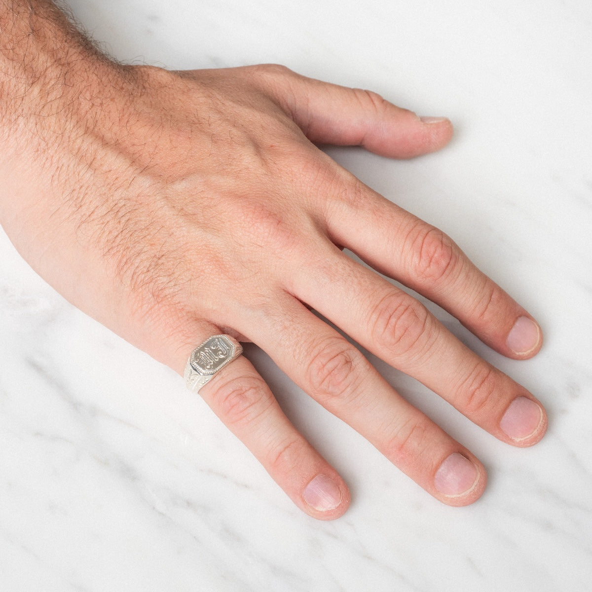 A close-up of a left hand on white marble, wearing the Sovereign Signet Ring—a silver ring with an engraved design on its flat, oval-shaped top.