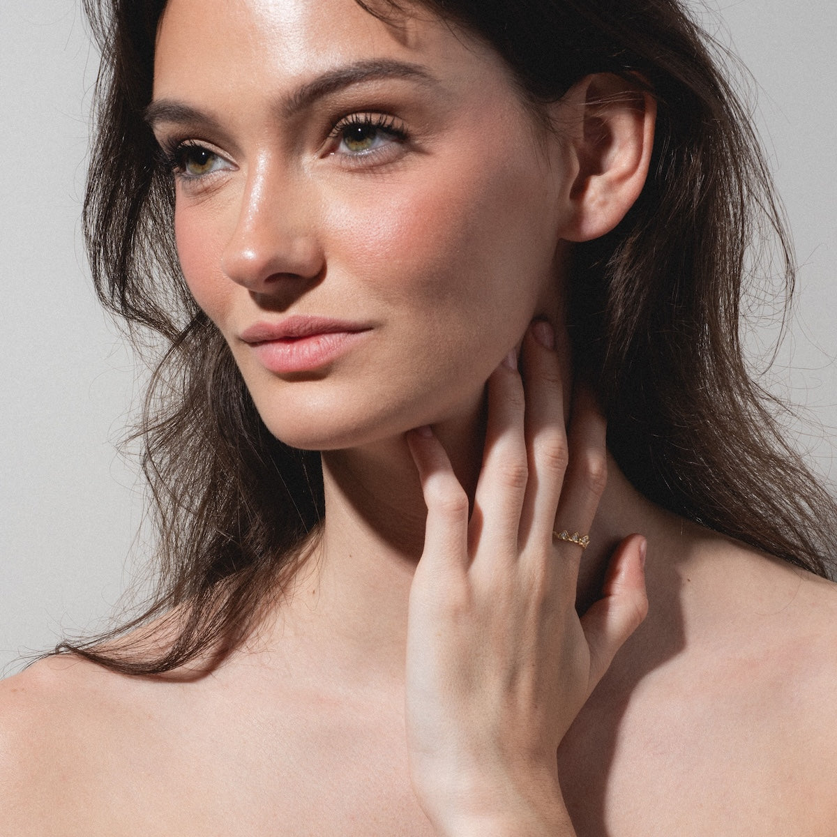 A woman with long brown hair and light eyes, wearing natural makeup, gazes to the side and gently touches her neck, showcasing the delicate Radiant Hearts Band (outward band) gold ring against a plain, neutral background.
