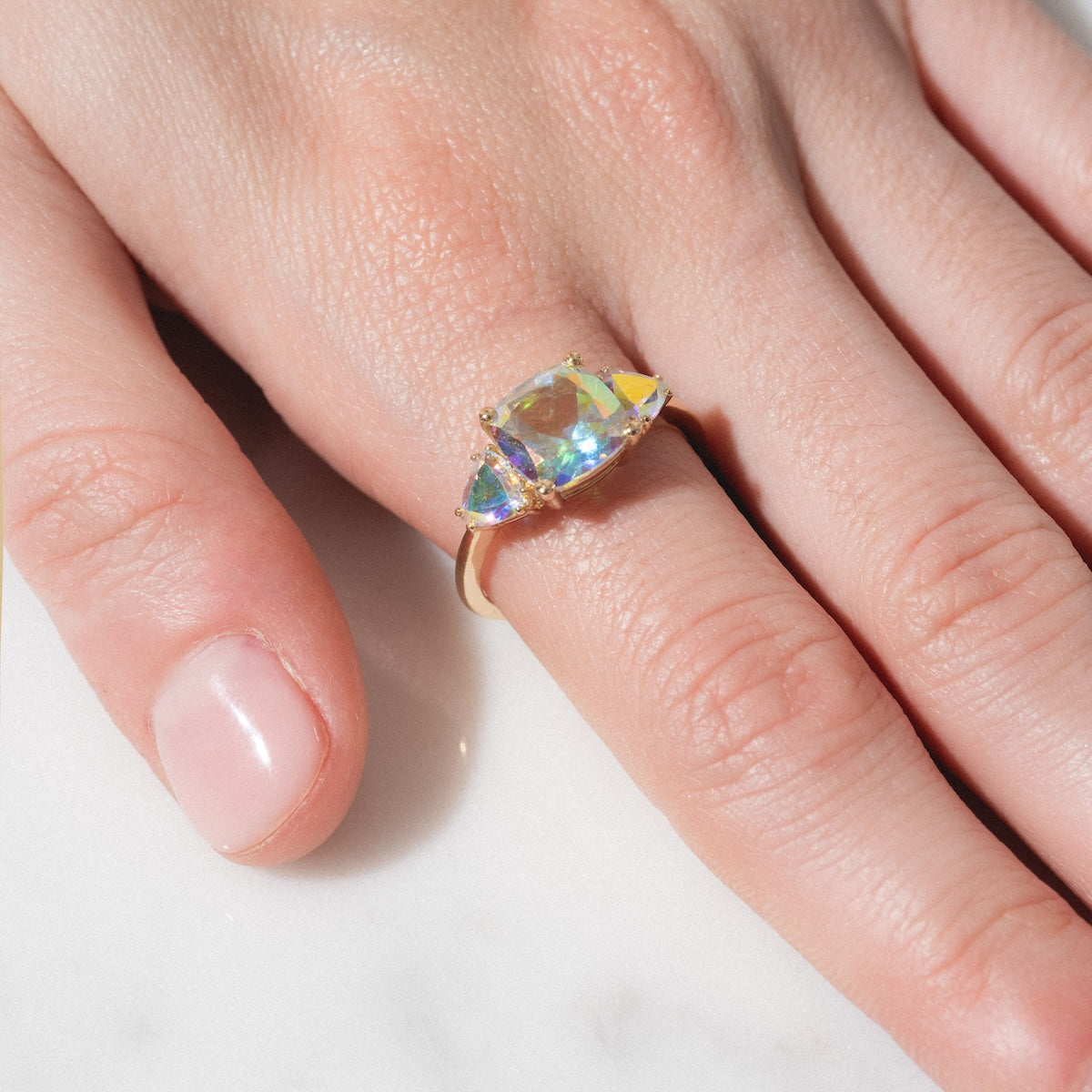 A hand with neatly manicured nails wears the Seraphina Ring—a gold band with a large iridescent gemstone flanked by two smaller stones—resting on a white marble surface.