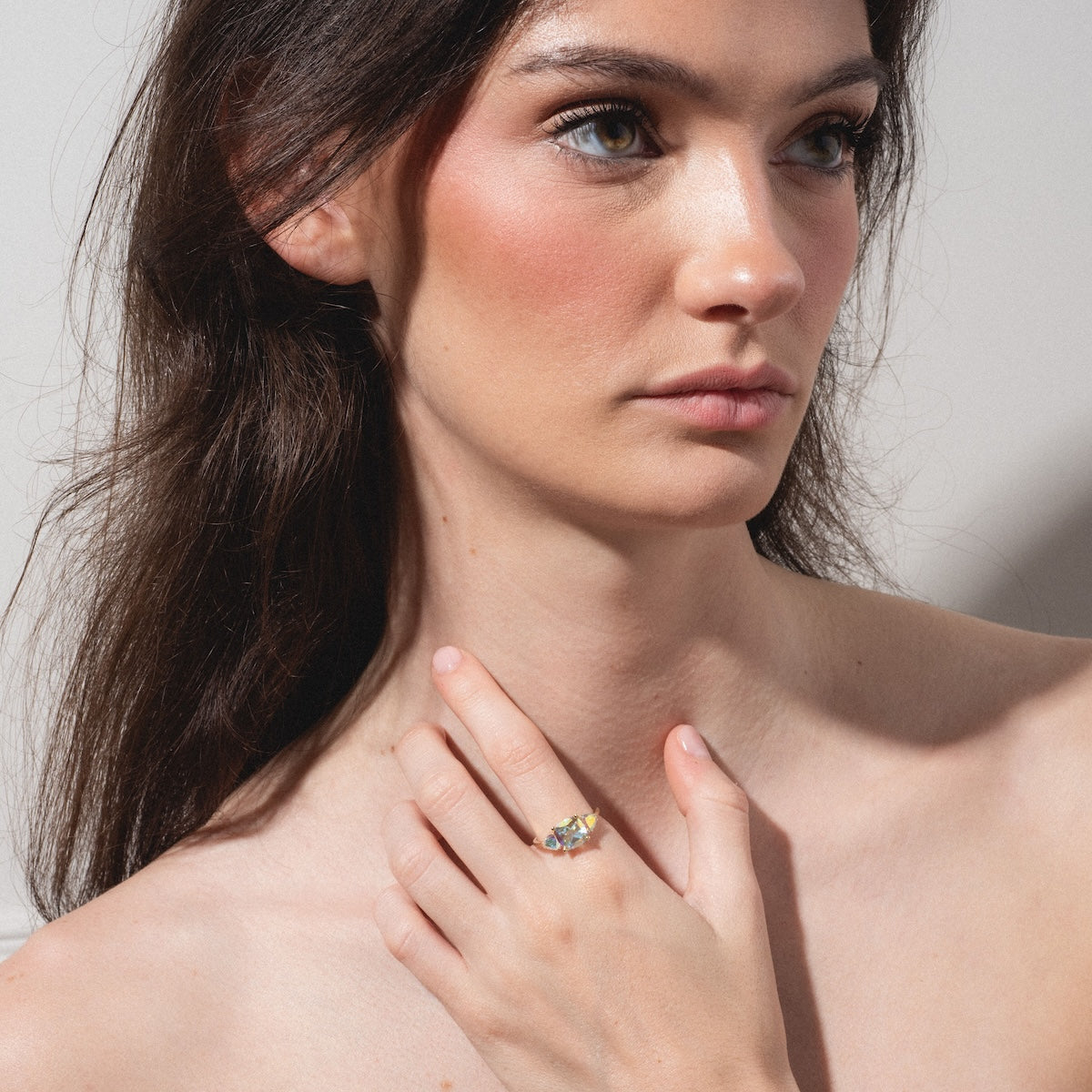 A woman with long brown hair and blue eyes gazes to the side, her left hand with the Seraphina Ring resting on her upper chest. She has a natural makeup look and bare shoulders.