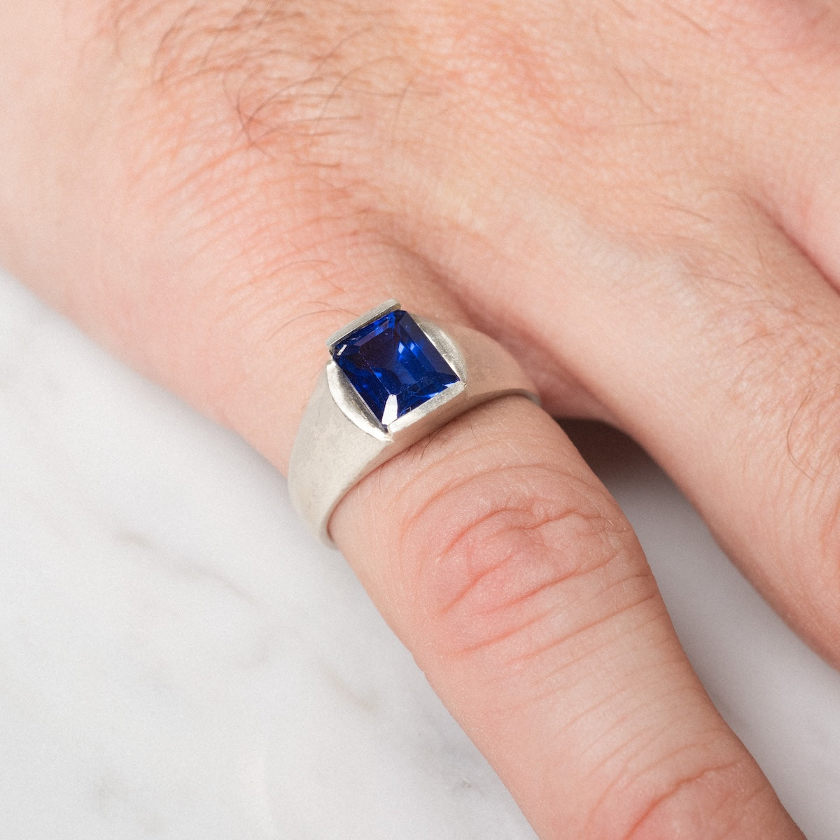 A hand wearing the Legacy Ring—a silver ring with a rectangular deep blue gemstone—on the ring finger, displayed against a light marble surface.