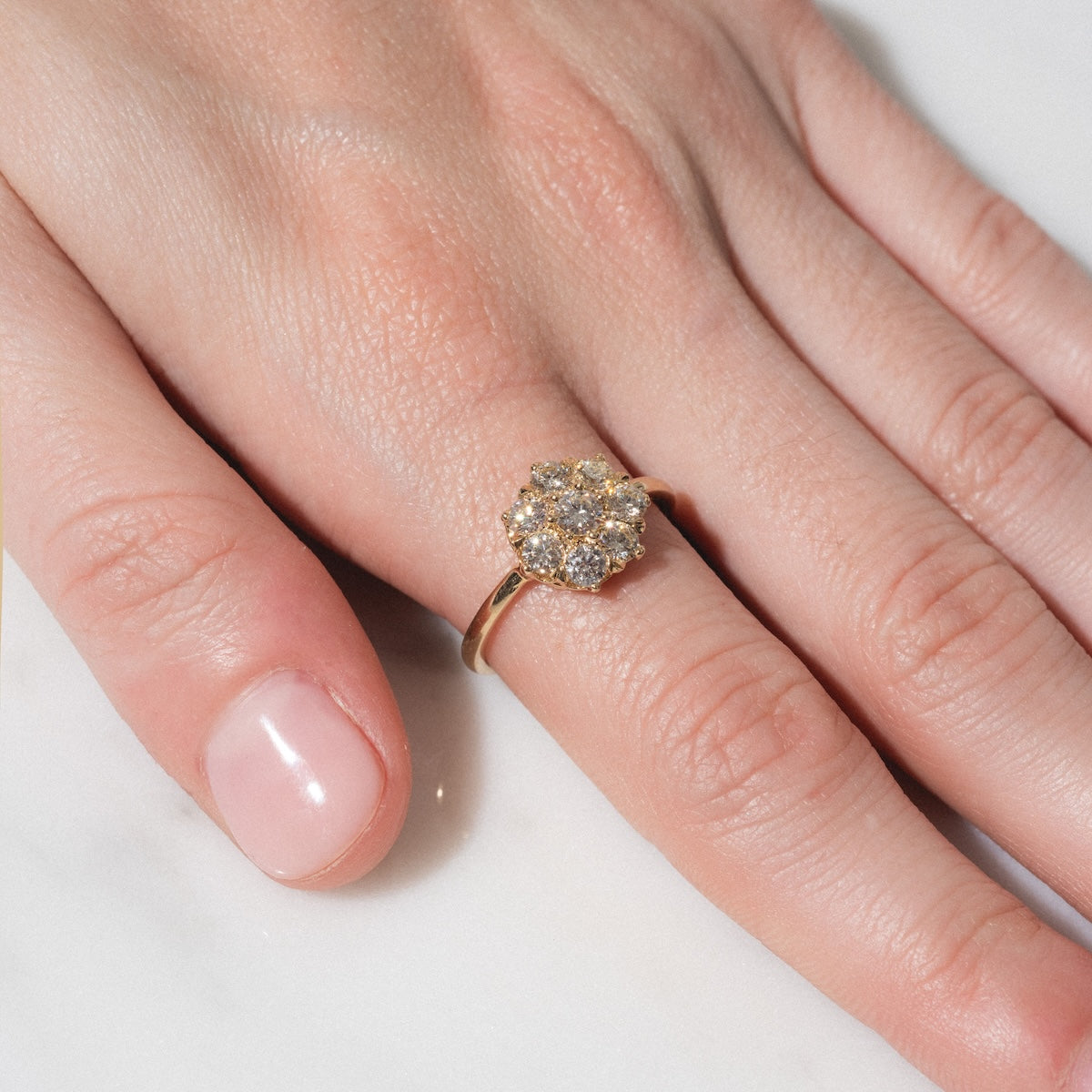 A hand rests on a white surface, wearing the Amas d'Etoiles Ring, featuring a gold band adorned with a cluster of sparkling diamonds in a floral pattern on the ring finger.