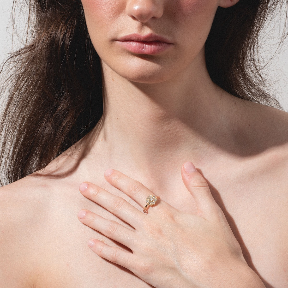 A woman with long brown hair and bare shoulders gently rests her hand on her chest, showcasing the Amas d'Etoiles Ring—a gold piece adorned with a sparkling cluster of stones—against a softly lit, plain background.