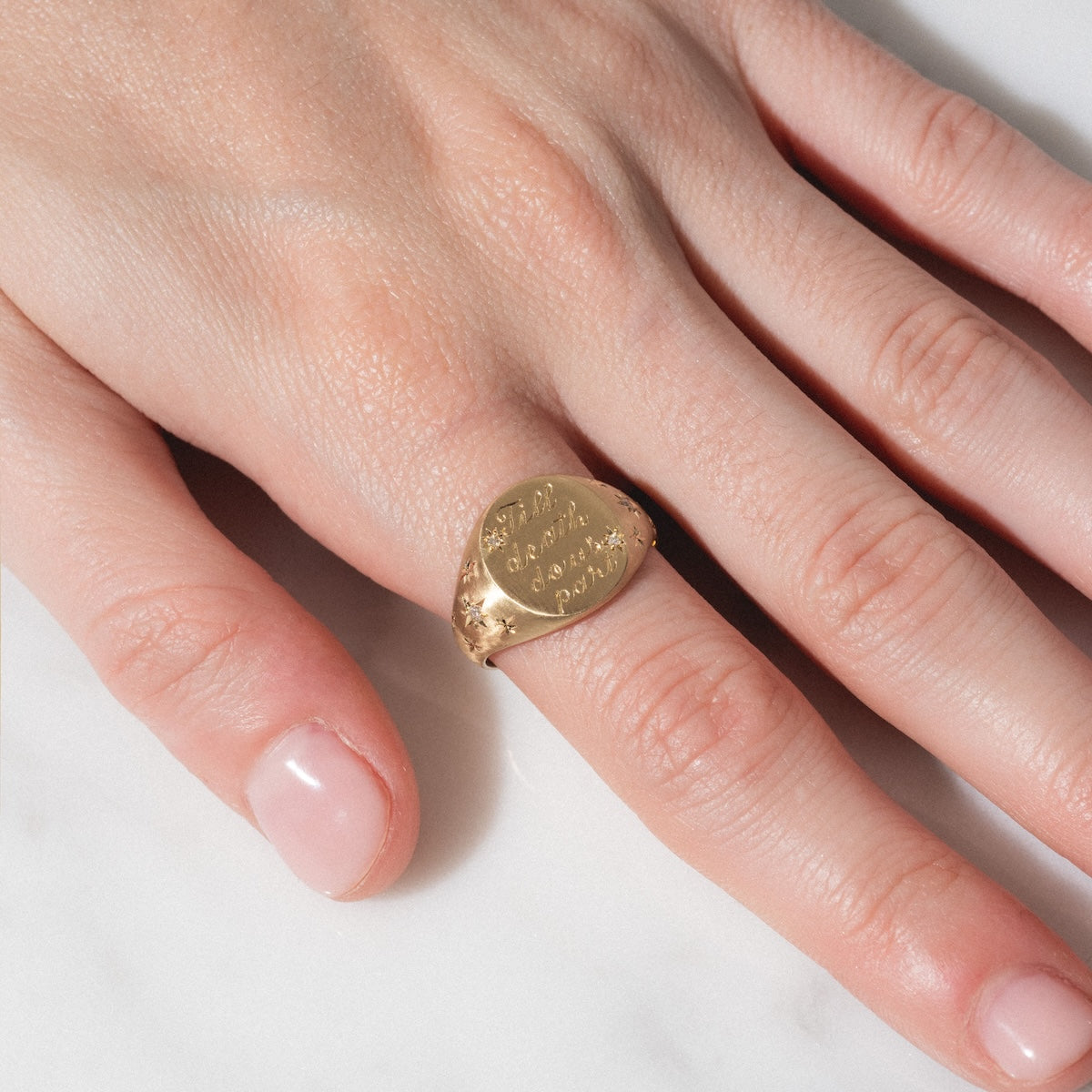 A close-up of a hand with neatly manicured nails wearing the Till Death Do Us Part Signet with Diamonds, resting on a white marble surface.