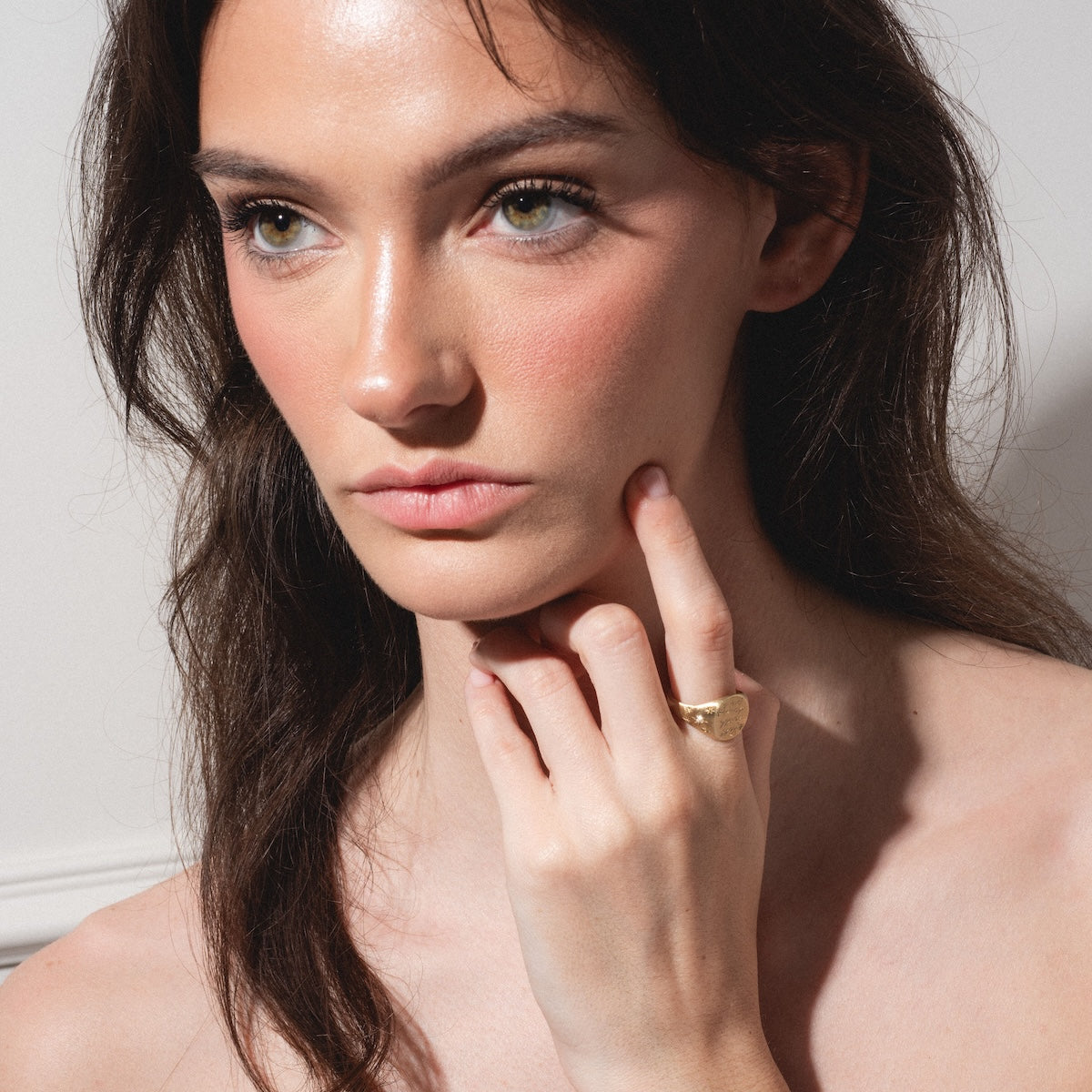 A woman with long brown hair and dewy makeup gazes thoughtfully to the side, touching her cheek. She wears the Till Death Do Us Part Signet with Diamonds on her finger against a softly lit, plain background.