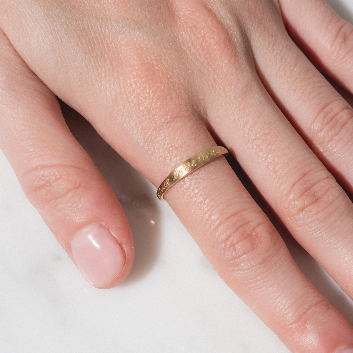 A close-up of a hand with natural, unpolished nails rests on a white surface, showcasing the True Love Never Dies Band in gold on the ring finger.