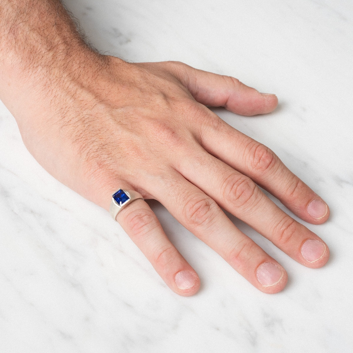 A hand rests on a white marble surface, wearing the Legacy Ring—a silver pinky ring featuring a rectangular blue gemstone.