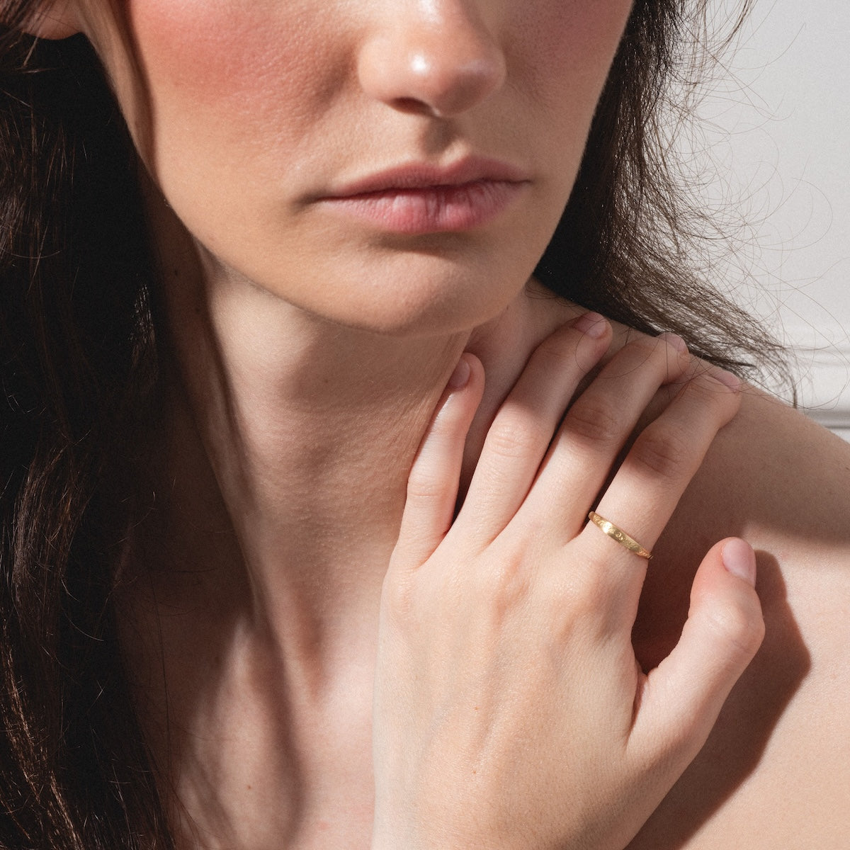 A woman with fair skin and brown hair rests her hand on her collarbone, displaying the True Love Never Dies Band in gold on her finger. Her parted lips and partially out-of-frame face add a touch of mystery to the scene.