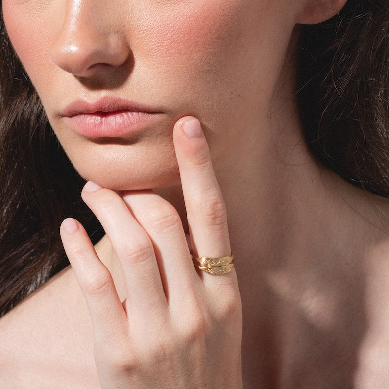 A woman with long brown hair and hazel eyes gazes thoughtfully to the side, her hand touching her chin as she wears the 14k Ripples Customizable ID Band and natural makeup with subtle pink blush.