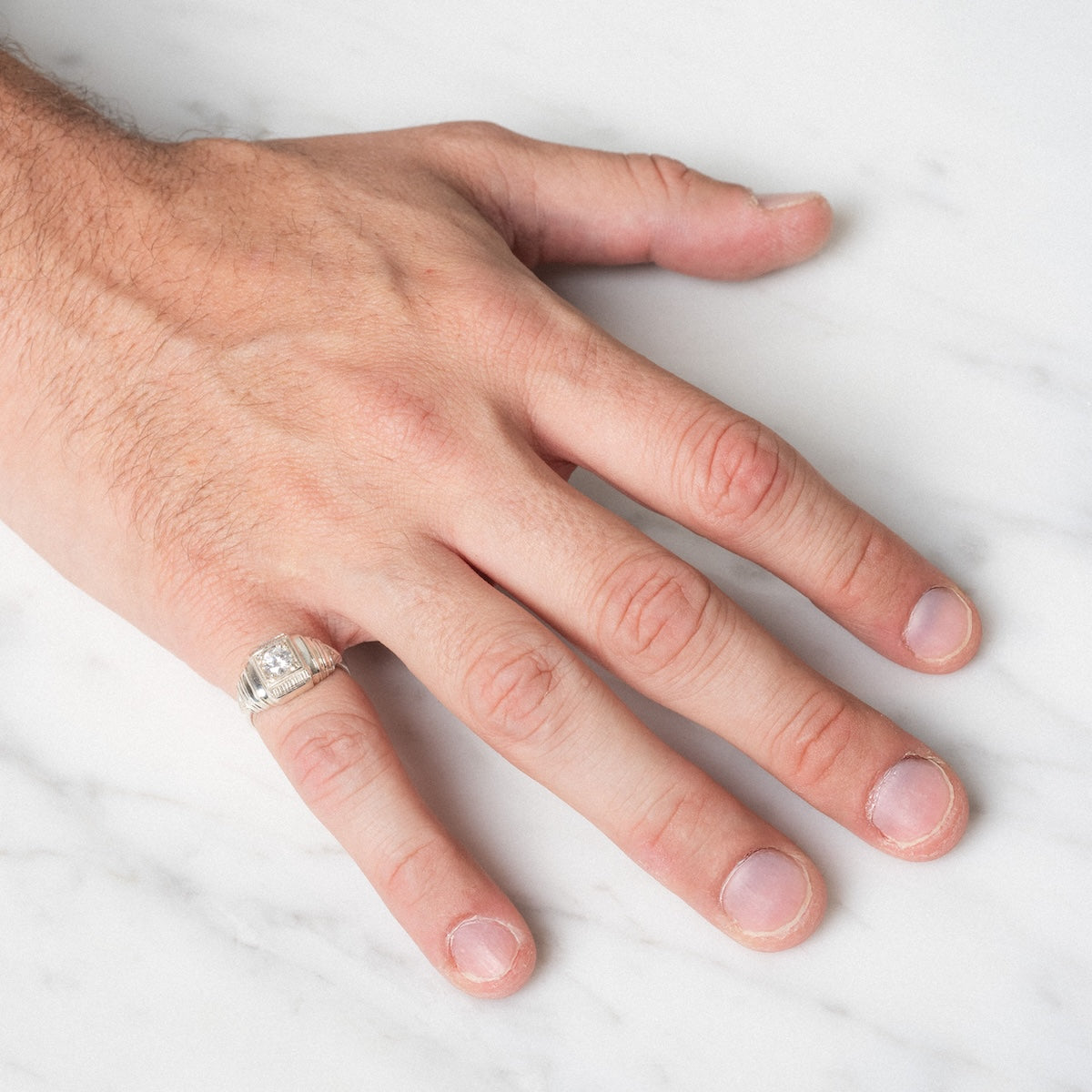 A person's left hand with short, slightly uneven nails rests on a white marble surface, wearing the Sacred Ring—a silver ring with a rectangular design—on the ring finger.