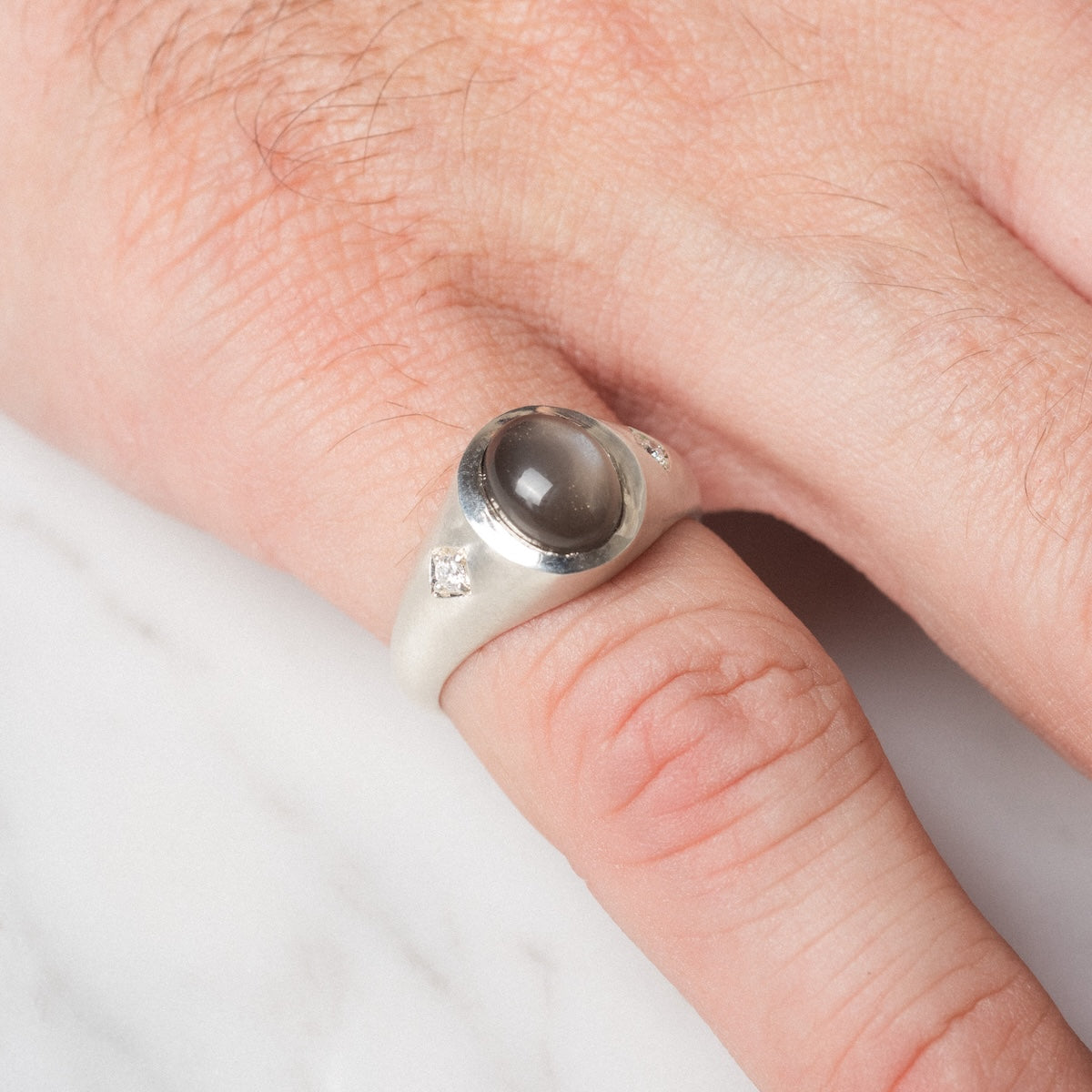 A close-up of a hand wearing the Vailance Ring, featuring a silver band with an oval gray center stone and two small clear stones on each side, resting on a white surface.