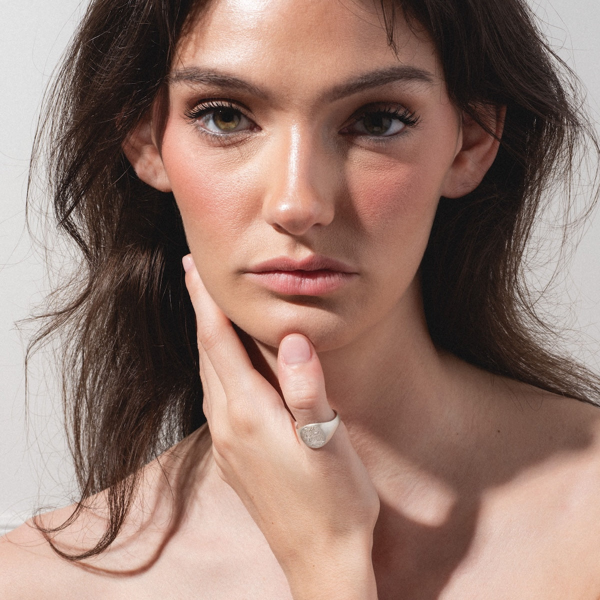 A woman with long brown hair and natural makeup gazes at the camera, holding her chin to display the Till Death Do Us Part Signet Ring on her finger against a plain, light-colored background.