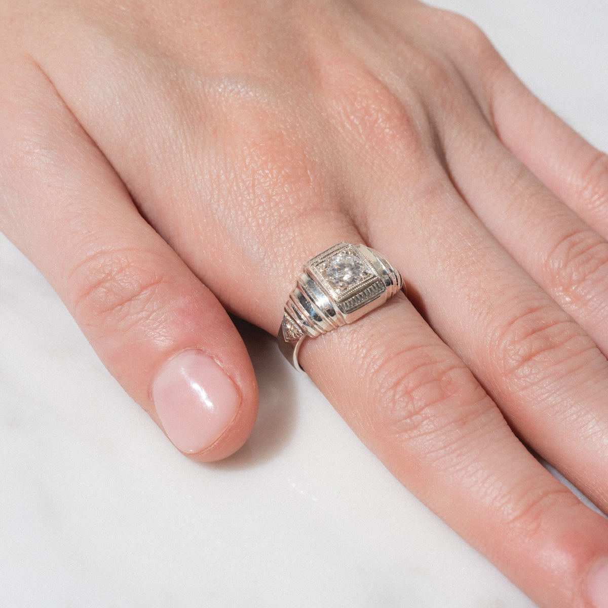 A close-up of a hand resting on a white surface, wearing the Sovereign Signet Ring—a silver ring featuring a large square-cut clear gemstone and decorative detailing.