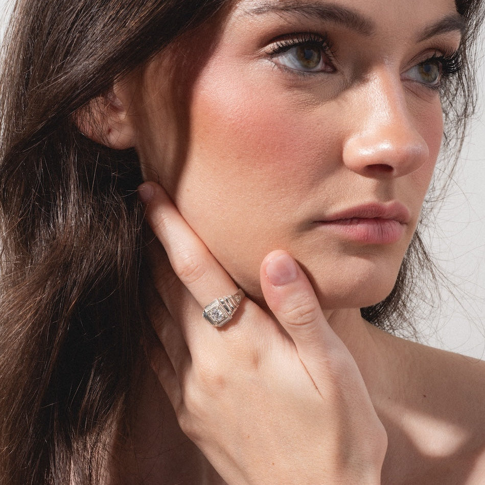 A woman with long brown hair gazes thoughtfully to the side, her hand resting on her neck to showcase the Sacred Ring—a detailed silver design—against a plain, light-colored background.