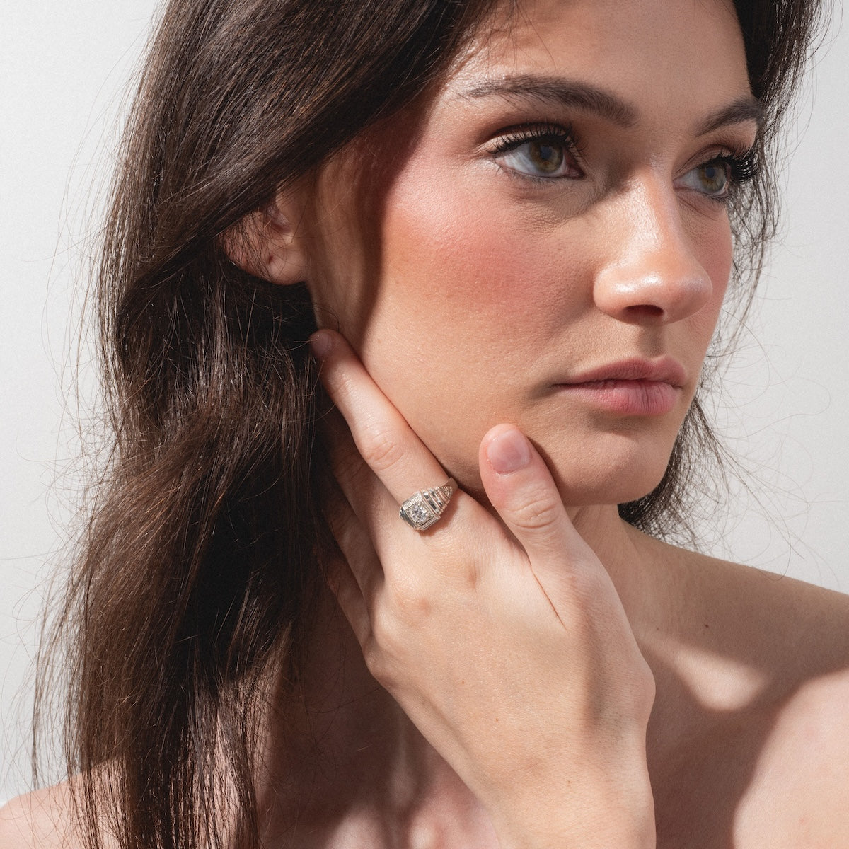 A woman with long brown hair gazes thoughtfully to the side, resting her chin on her hand. She wears the Sovereign Signet Ring on her finger, complemented by natural makeup, soft blush, and defined brows against a plain light background.