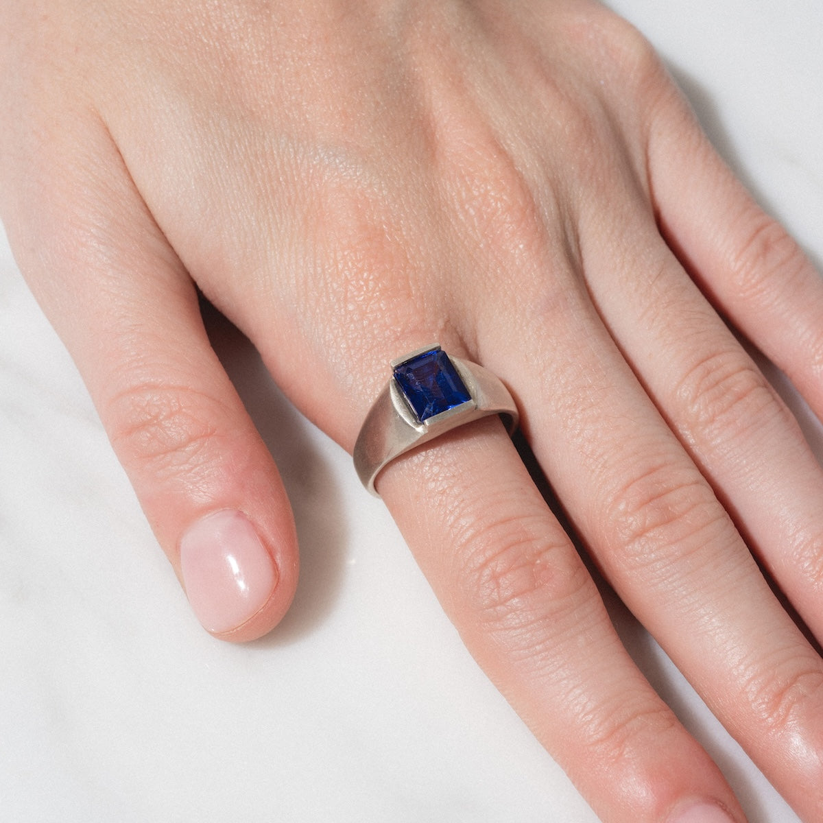 A hand with neatly manicured nails wears the Legacy Ring, featuring a rectangular blue gemstone set in silver, displayed on a light-colored surface.