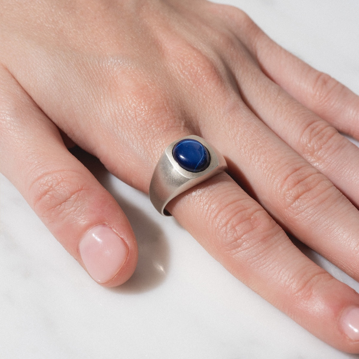 A hand rests on a white surface, wearing the Guardian Ring—a silver band featuring a smooth, oval dark blue stone—on the middle finger.