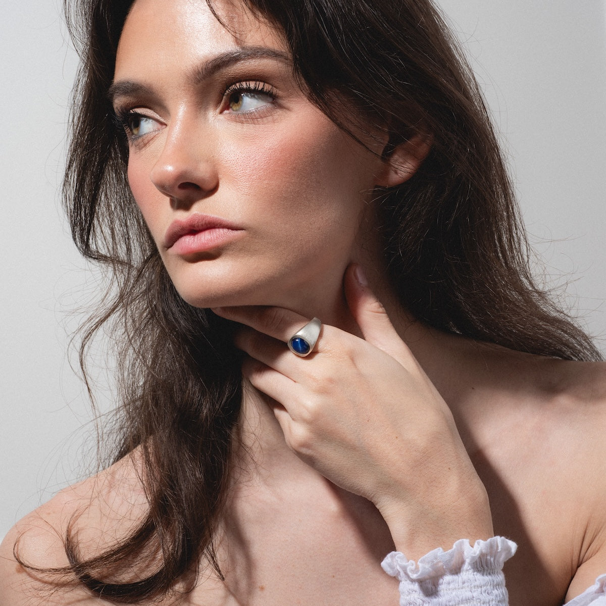A woman with long brown hair gazes thoughtfully to the side, wearing a white off-shoulder top and the Guardian Ring—a large silver ring with a blue stone—on her hand resting gently on her neck.