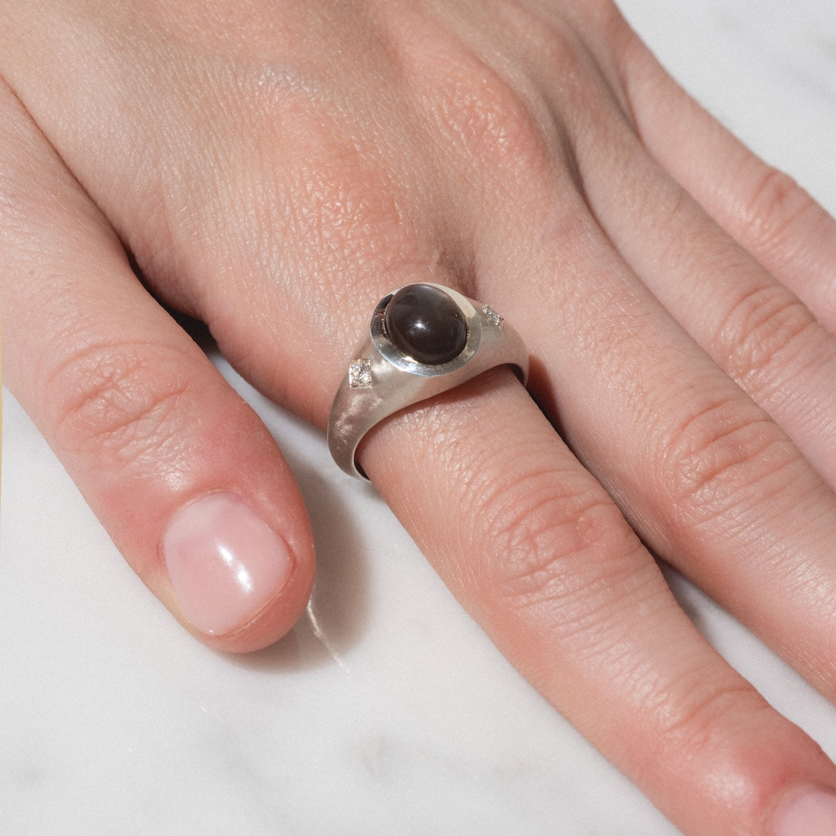 A close-up of a hand on a white surface, wearing the Vailance Ring—a silver band featuring a dark oval stone at its center.