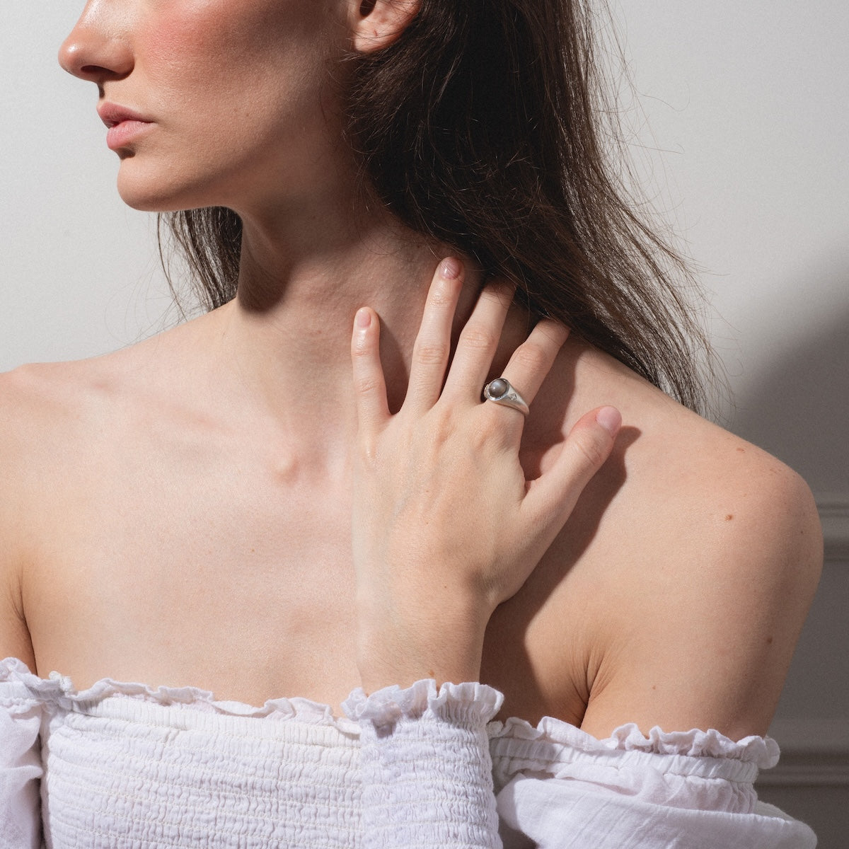 A woman in an off-shoulder white top touches her neck with her left hand, showcasing the Vailance Ring. Her face is partially visible, softly illuminated against a plain background.