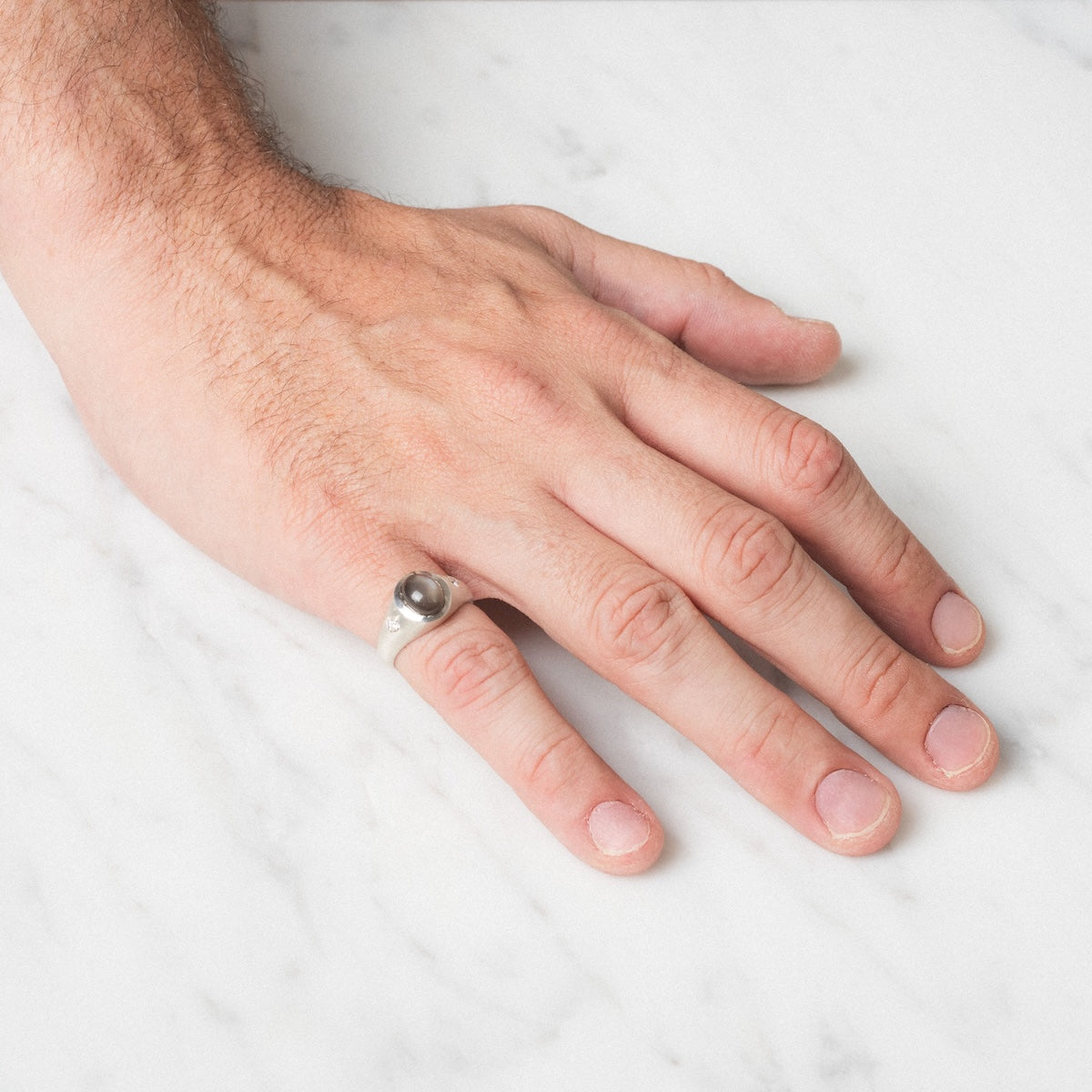A hand rests on a white marble surface, wearing the Vailance Ring—a silver band featuring a round, dark stone on the ring finger.