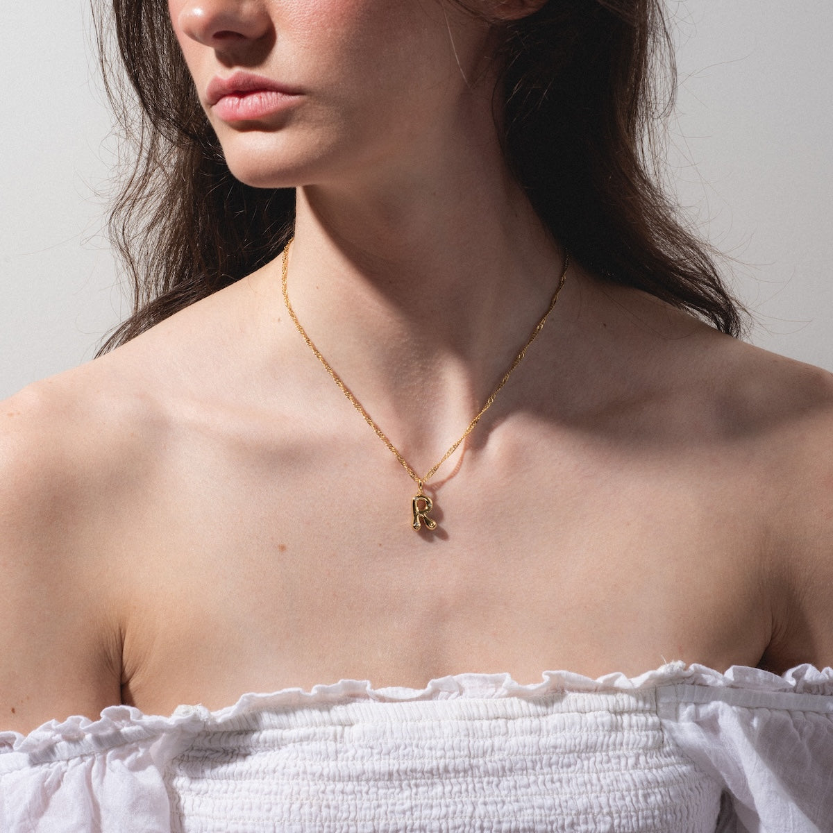 A woman in an off-the-shoulder white top wears the Star Crossed Celestial Bubble Letter Charm gold necklace with an "R" pendant. Her face is turned partly away from the camera.