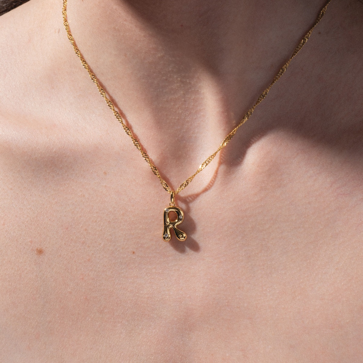 A close-up shows a fair-skinned person’s neck wearing the Star Crossed Celestial Bubble Letter Charm— a gold chain necklace with a gold letter R pendant; no clothing is visible.