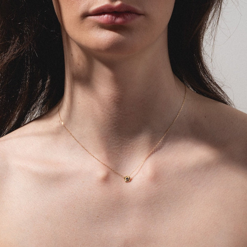 A woman wears a Solid Gold Birth Flower Bloom Necklace with a small pendant and an off-the-shoulder white top, photographed from the shoulders up against a neutral background.