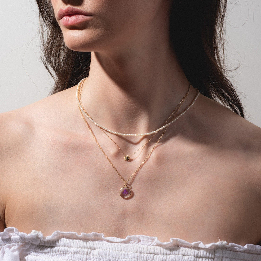 A woman wears an off-shoulder white top and three gold necklaces, including the Solid Gold Birth Flower Bloom Necklace with a round purple gemstone. One necklace has a small letter pendant, another features a pearl-like strand. Her face is partially out of frame.