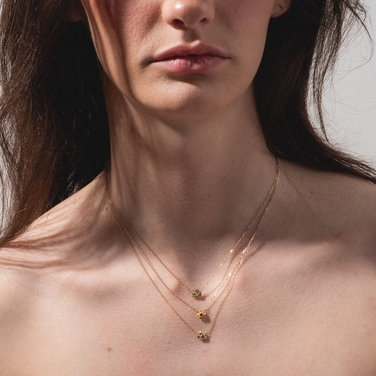 A woman with long dark hair wears three Solid Gold Birth Flower Bloom Necklaces of different lengths, each featuring a small pendant. Her face is partially visible and she poses against a plain background with a neutral expression.