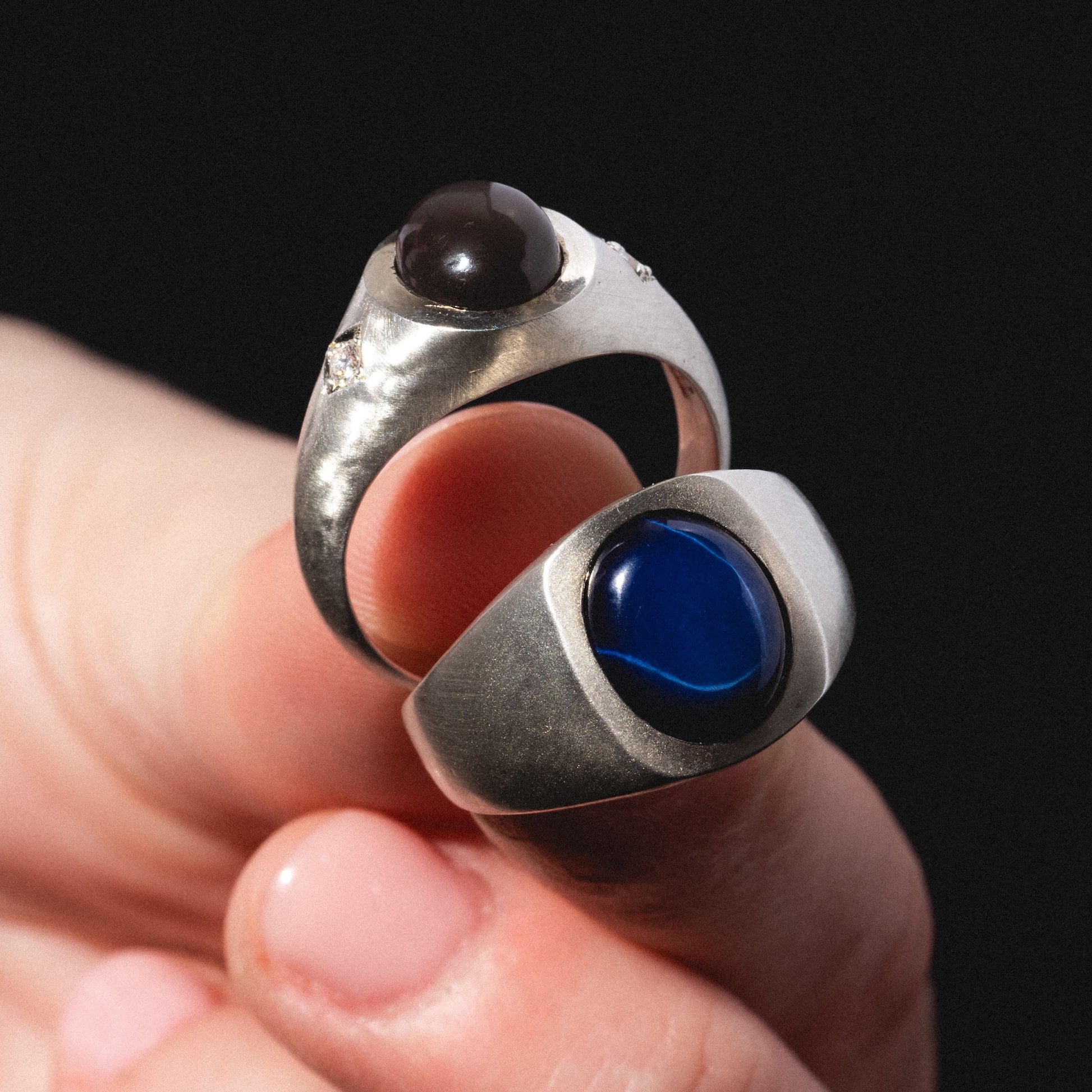 A hand displays two Vailance Rings—one featuring a dark brown cabochon stone, the other a deep blue oval stone—set against a black background.