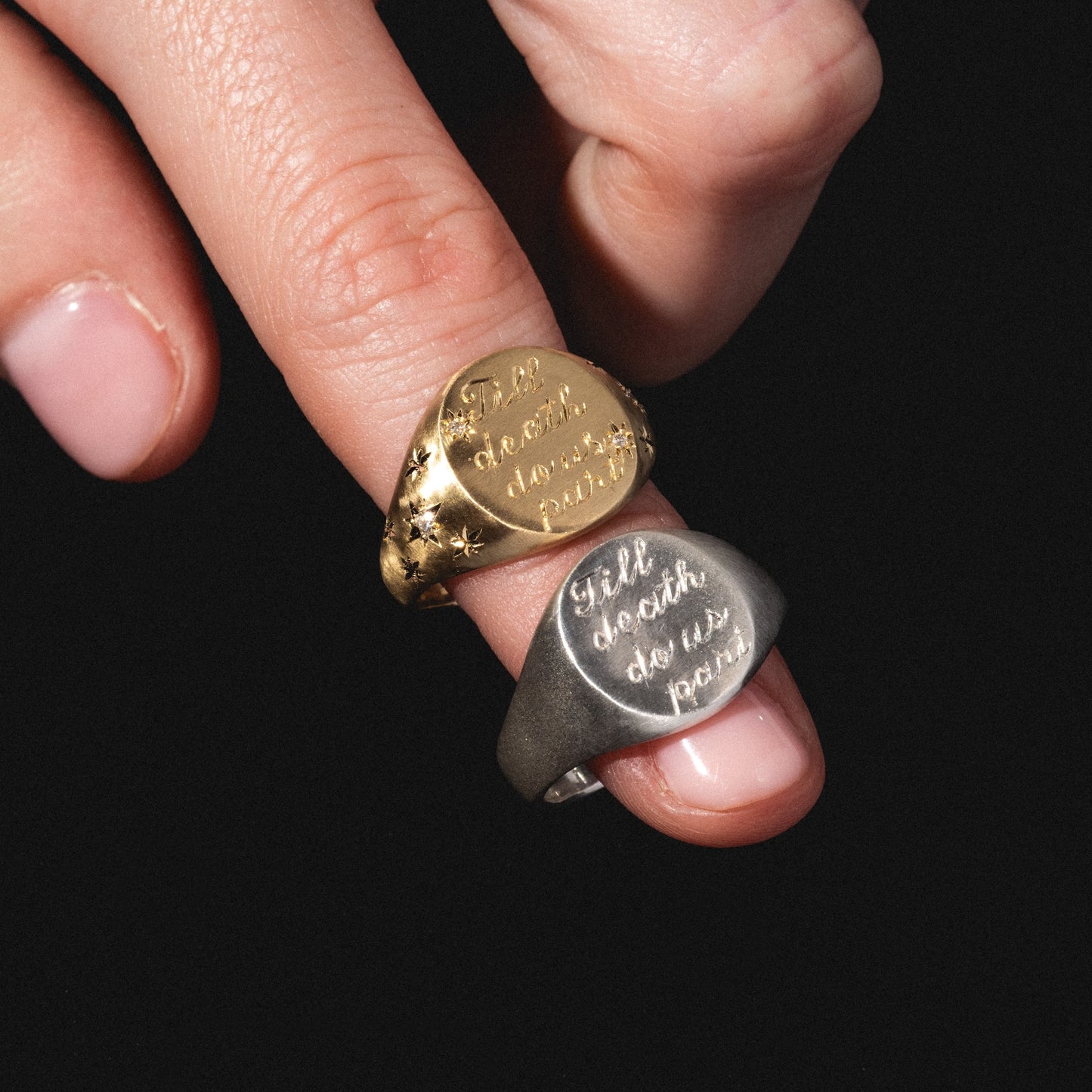 A hand with light pink nails holds two Till Death Do Us Part Signet rings with diamonds—one gold, one silver—against a black background.